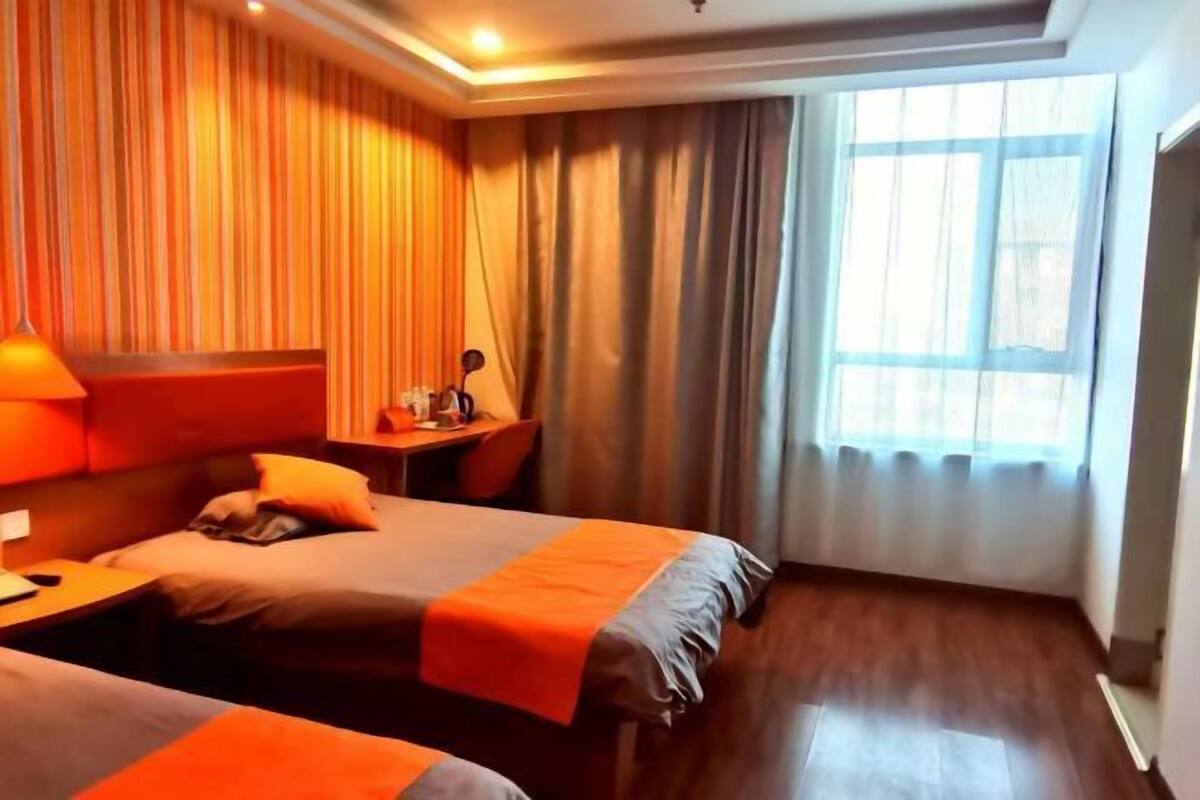 Habitación de hotel con dos camas, un escritorio, una silla, una lámpara y un ventanal con cortinas.
