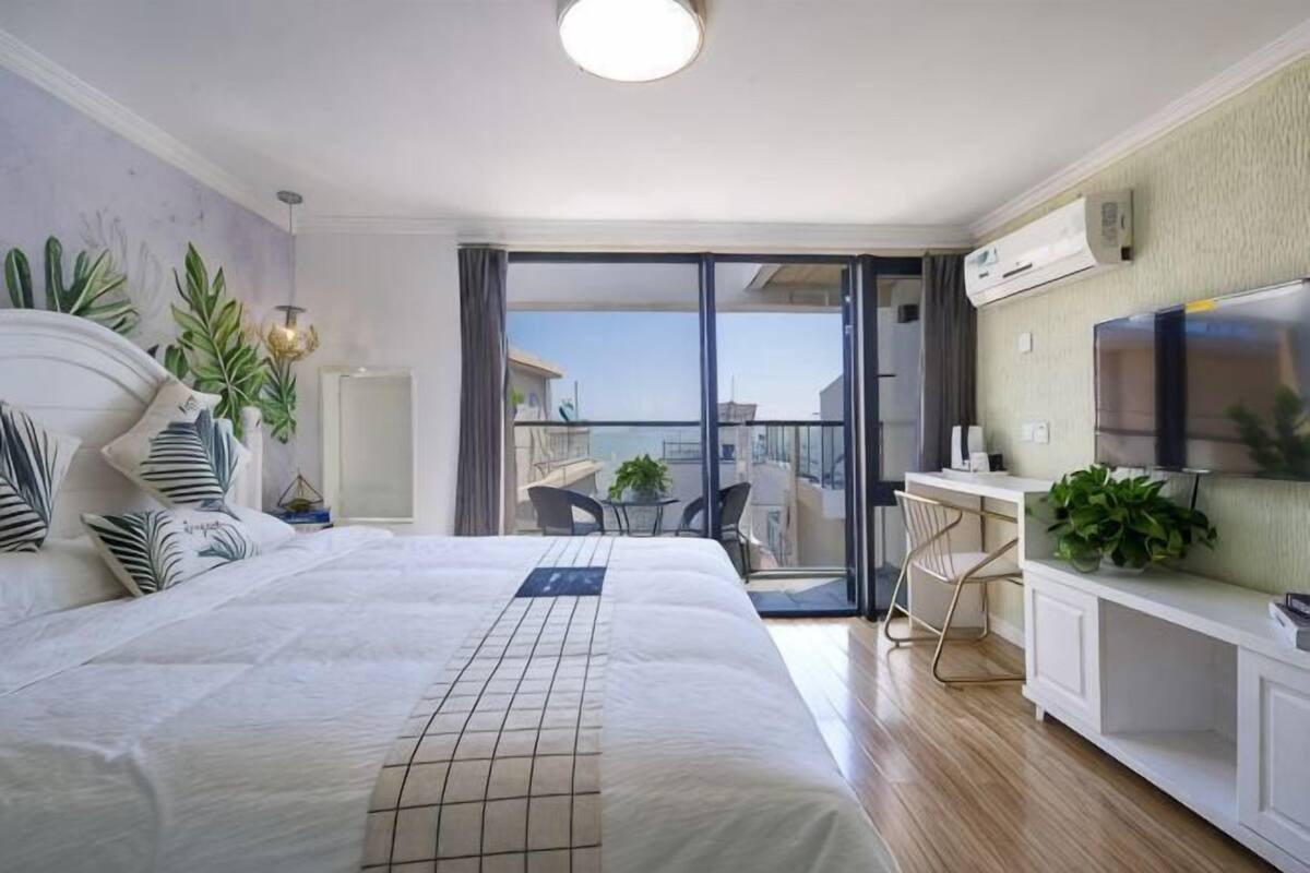A hotel room with a large bed, a balcony with a view, a TV, and a desk with a chair.