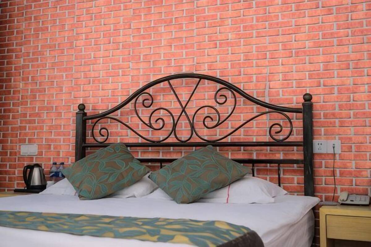 Un lit avec une tête de lit en métal, deux coussins à motifs, une table de chevet avec une lampe et un mur en briques.