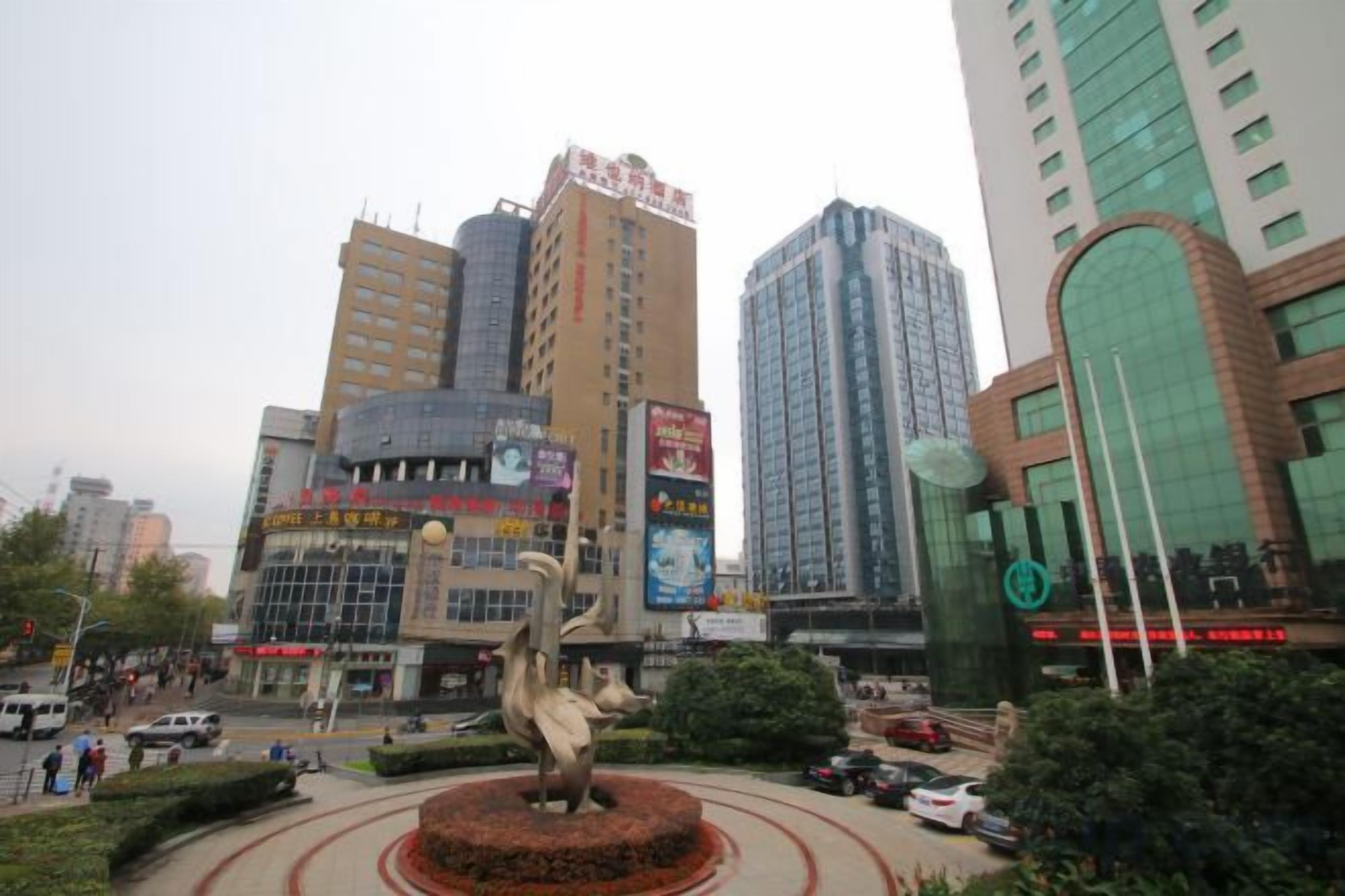 A cityscape with a central roundabout, a prominent sculpture, and tall buildings with commercial signage.