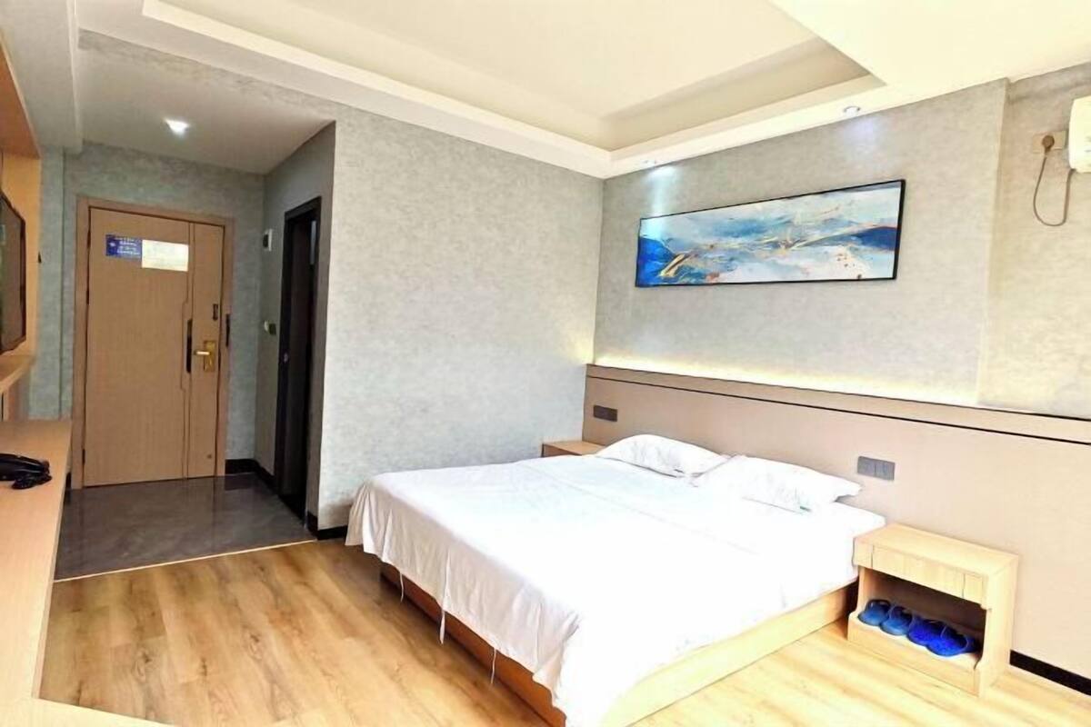 A hotel room with a bed, a small bedside table, a wall-mounted painting, and a door leading to another room.