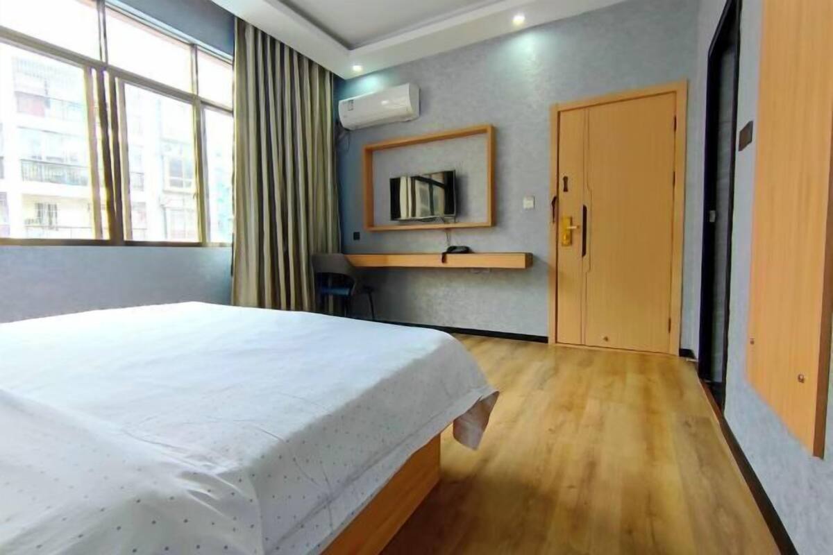 A hotel room with a bed, a TV on a wall, a wooden wardrobe, and a window with curtains.