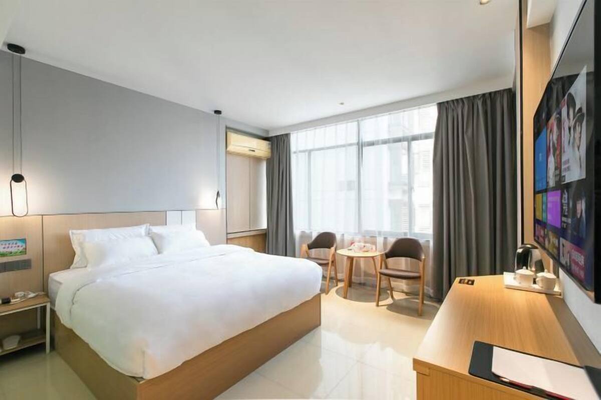 Habitació d'hotel amb un llit gran, un escriptori amb cadira, una taula petita amb caires i una finestra ampla amb cortines.