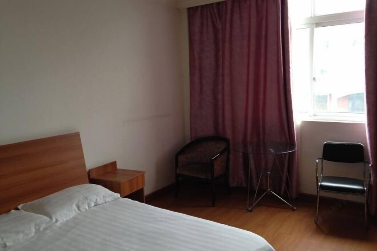 A hotel room with a wooden bed, a small wooden nightstand, a chair, a desk, and a window with curtains.