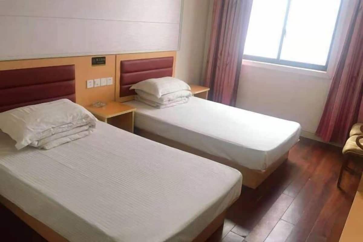 A hotel room with two single beds, wooden headboards, a small table, and a window with curtains.