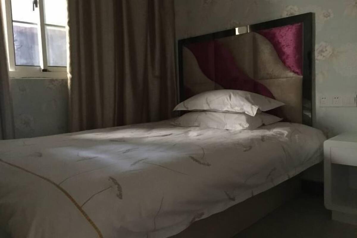 A single bed with a headboard, a window with curtains, and a white bedside table.