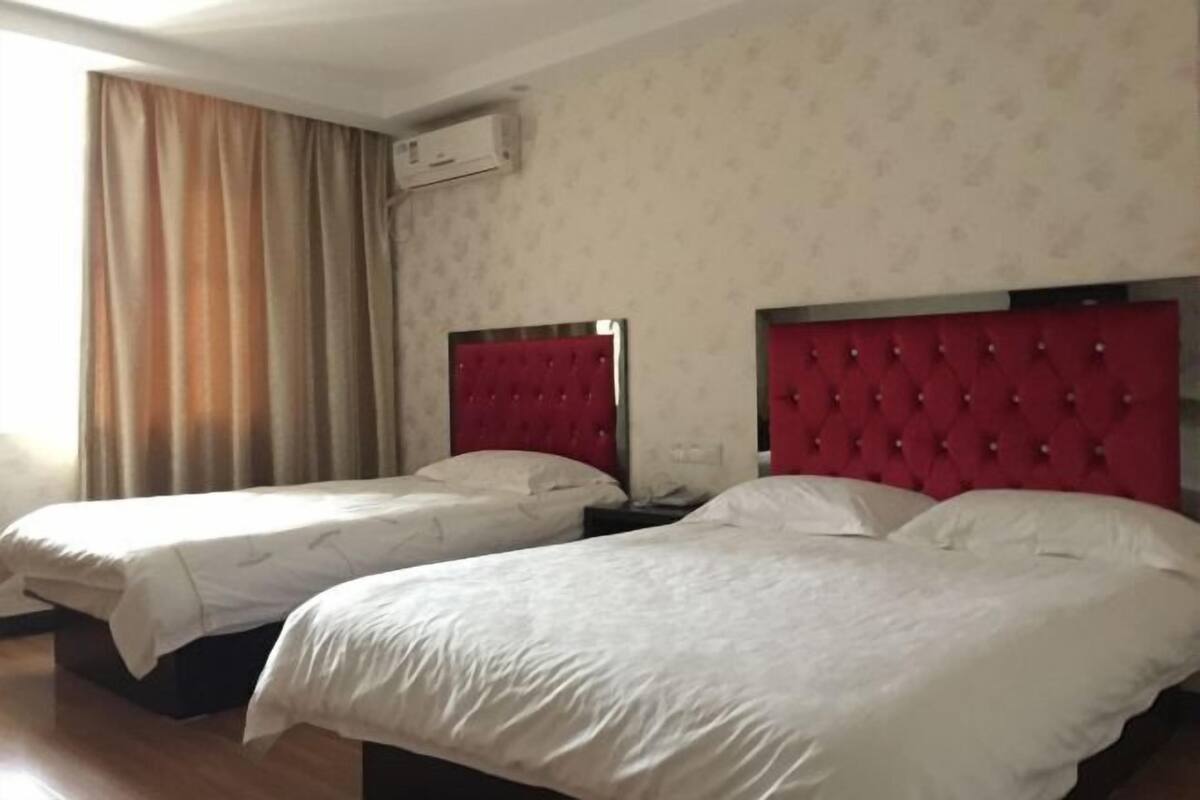 A hotel room with two beds, each with a red headboard, and a window with beige curtains.
