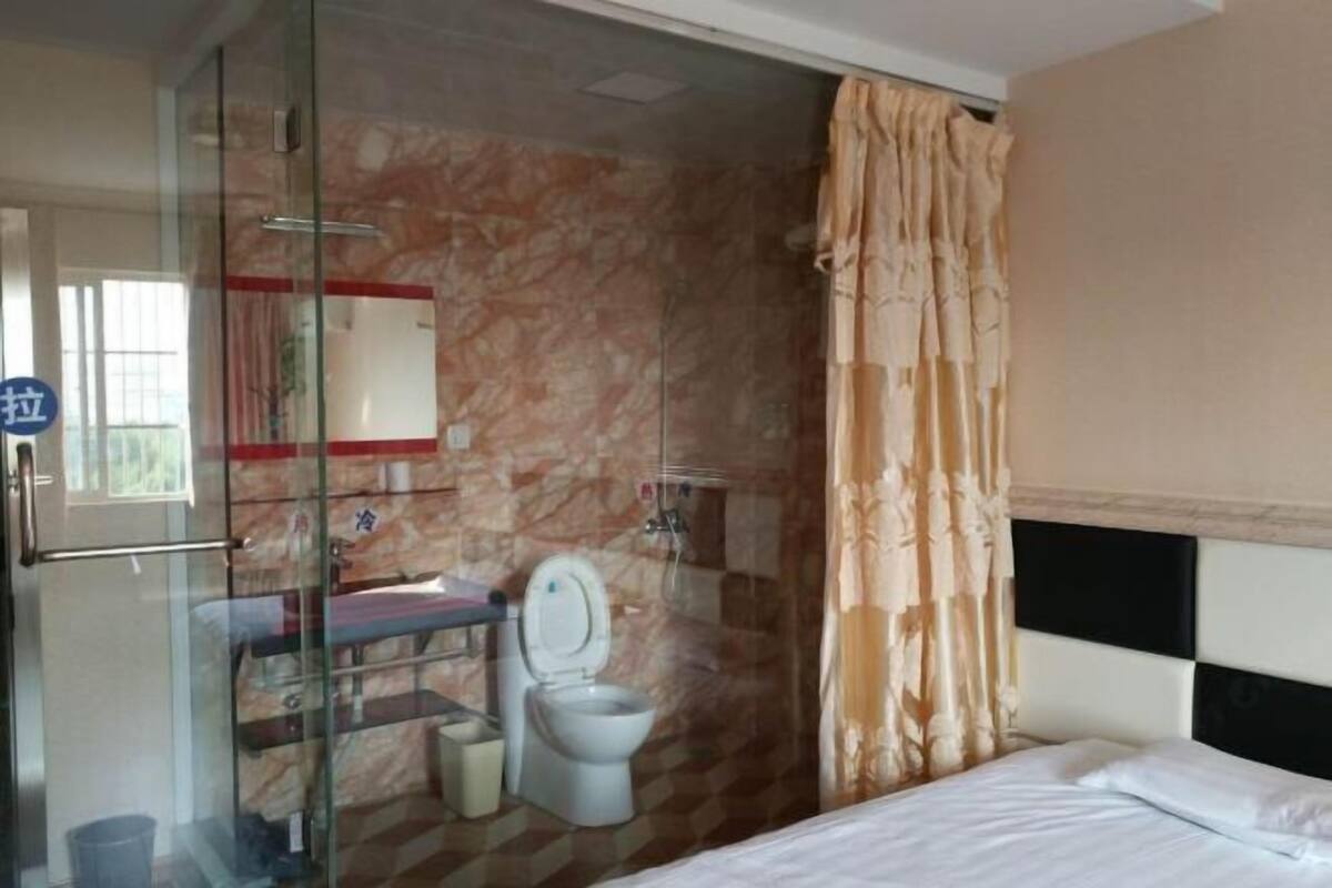 A hotel room with a glass shower enclosure, a marble wall, a bed with white bedding, and a bathroom with a toilet and sink.