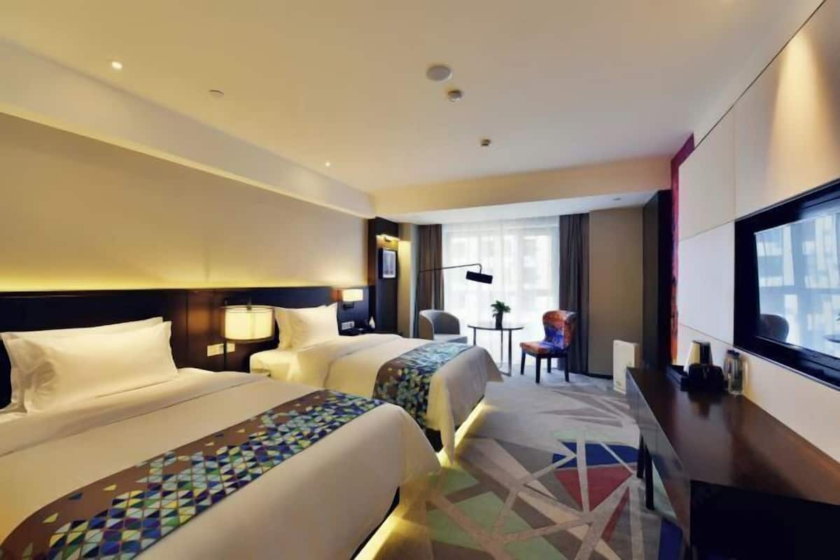 A hotel room with two beds, a desk, a chair, a TV, and a view of the city.