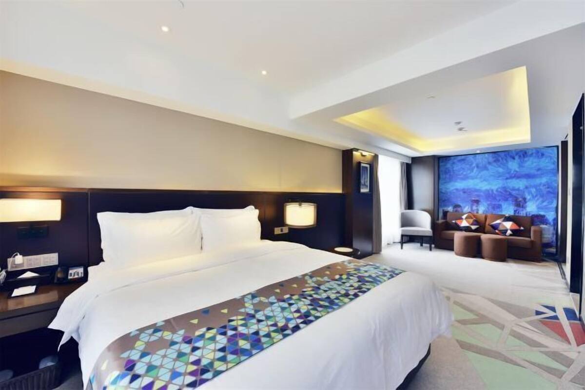 A modern hotel room with a large bed, a desk with a lamp, a seating area with a couch and chairs, and a large window with a cityscape view.