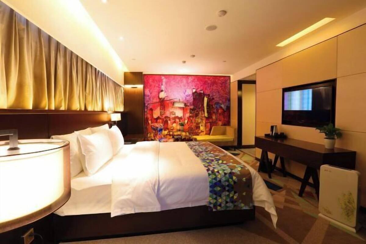 A hotel room with a bed, a desk, a TV, and a large abstract painting on the wall.