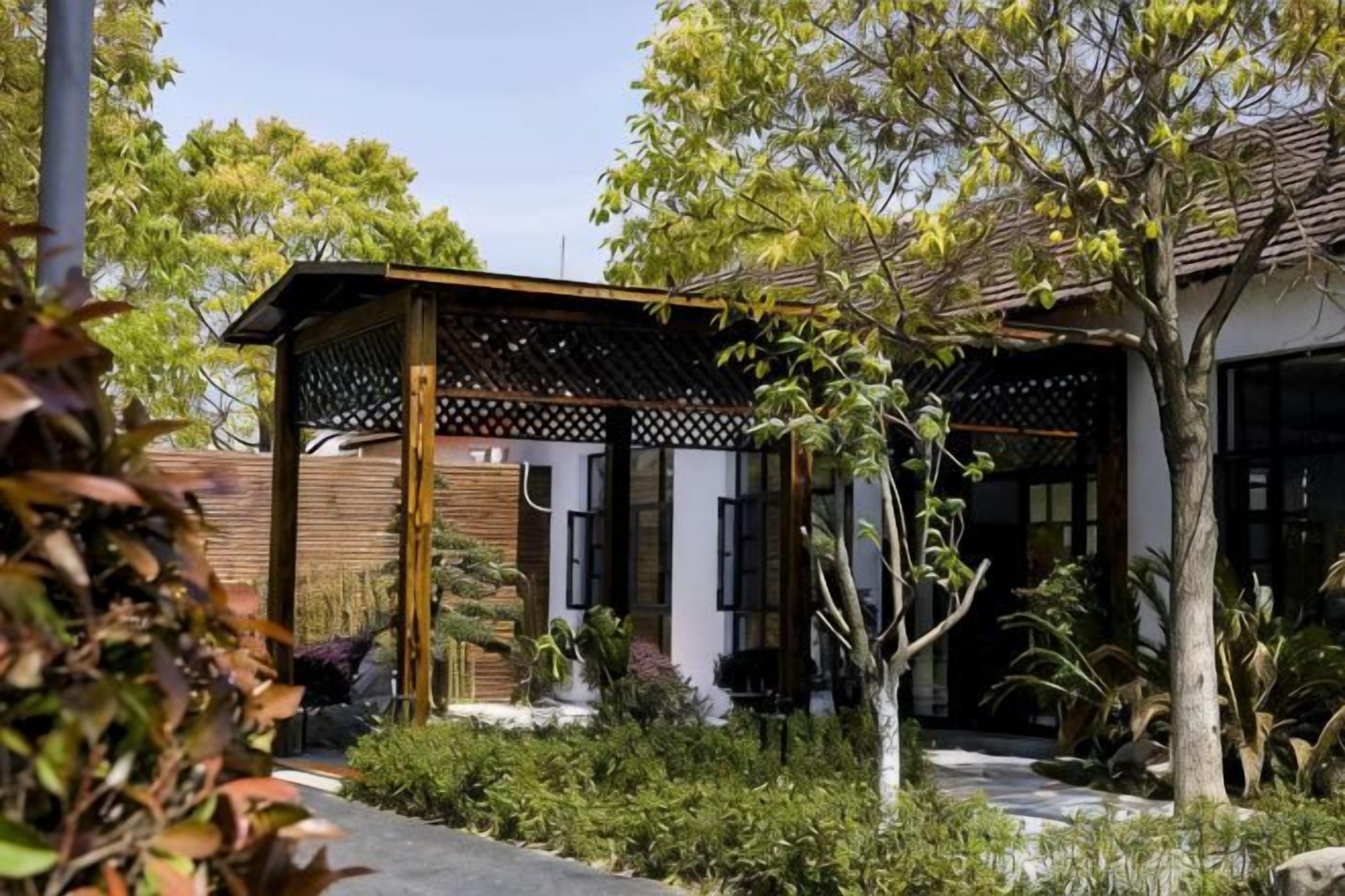A covered outdoor area with a wooden structure, surrounded by greenery and a pathway leading to a building with a tiled roof.
