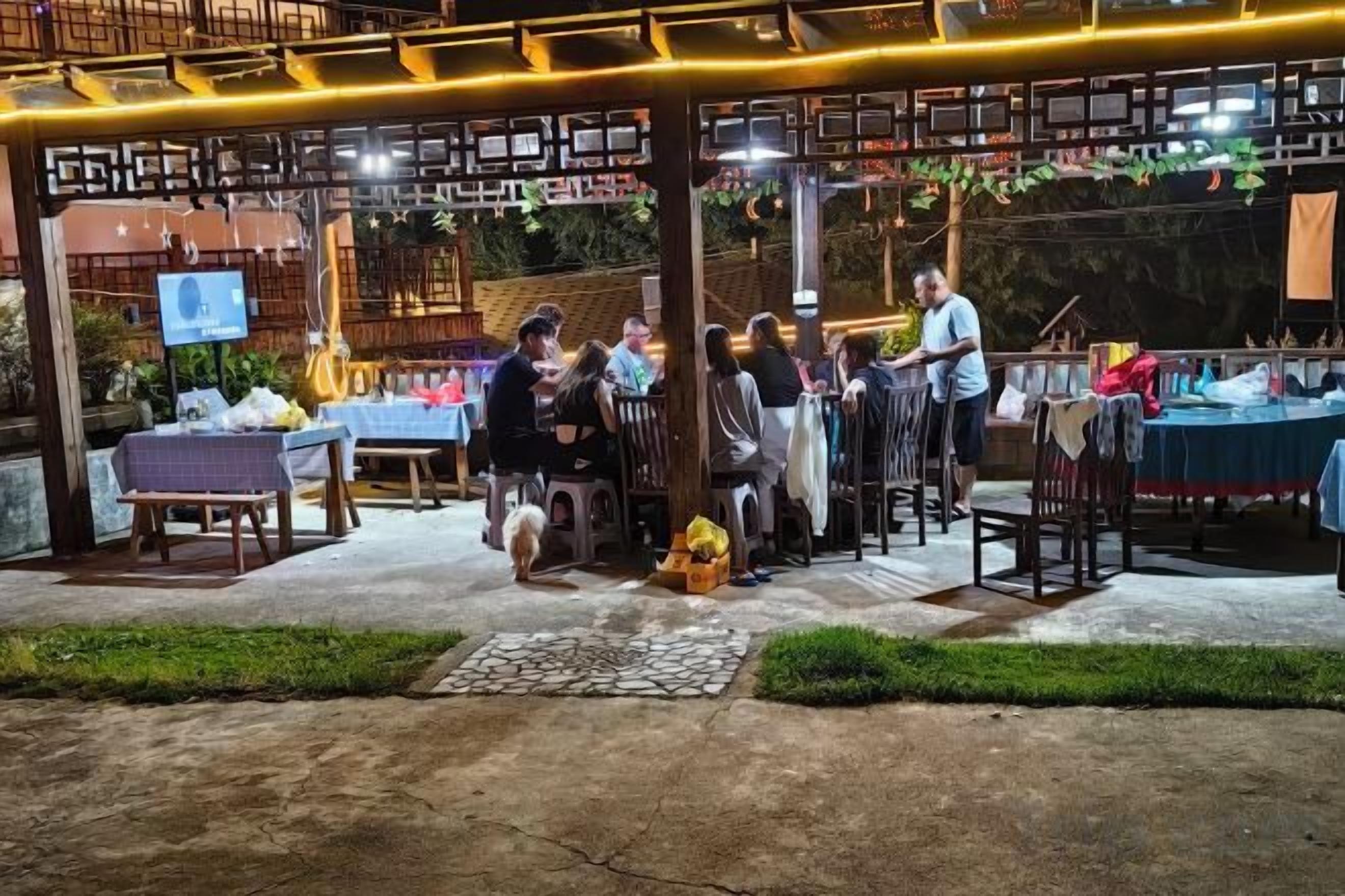 An outdoor dining area with tables and chairs, a wooden pergola, and a TV screen.