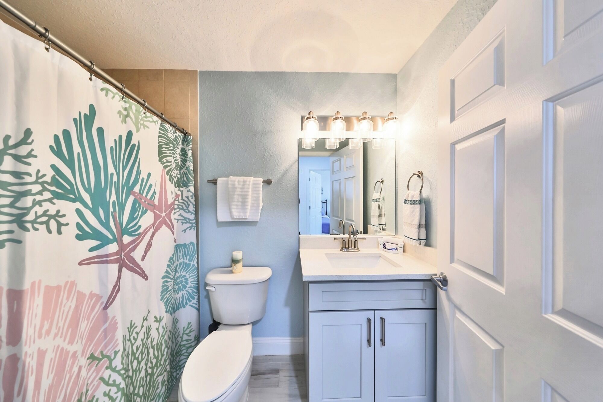 Et badeværelse med bruseforhæng med et tropisk blad- og stjerneskudsdesign, et hvidt toilet, en vask med et spejl over og et skab med to låger.