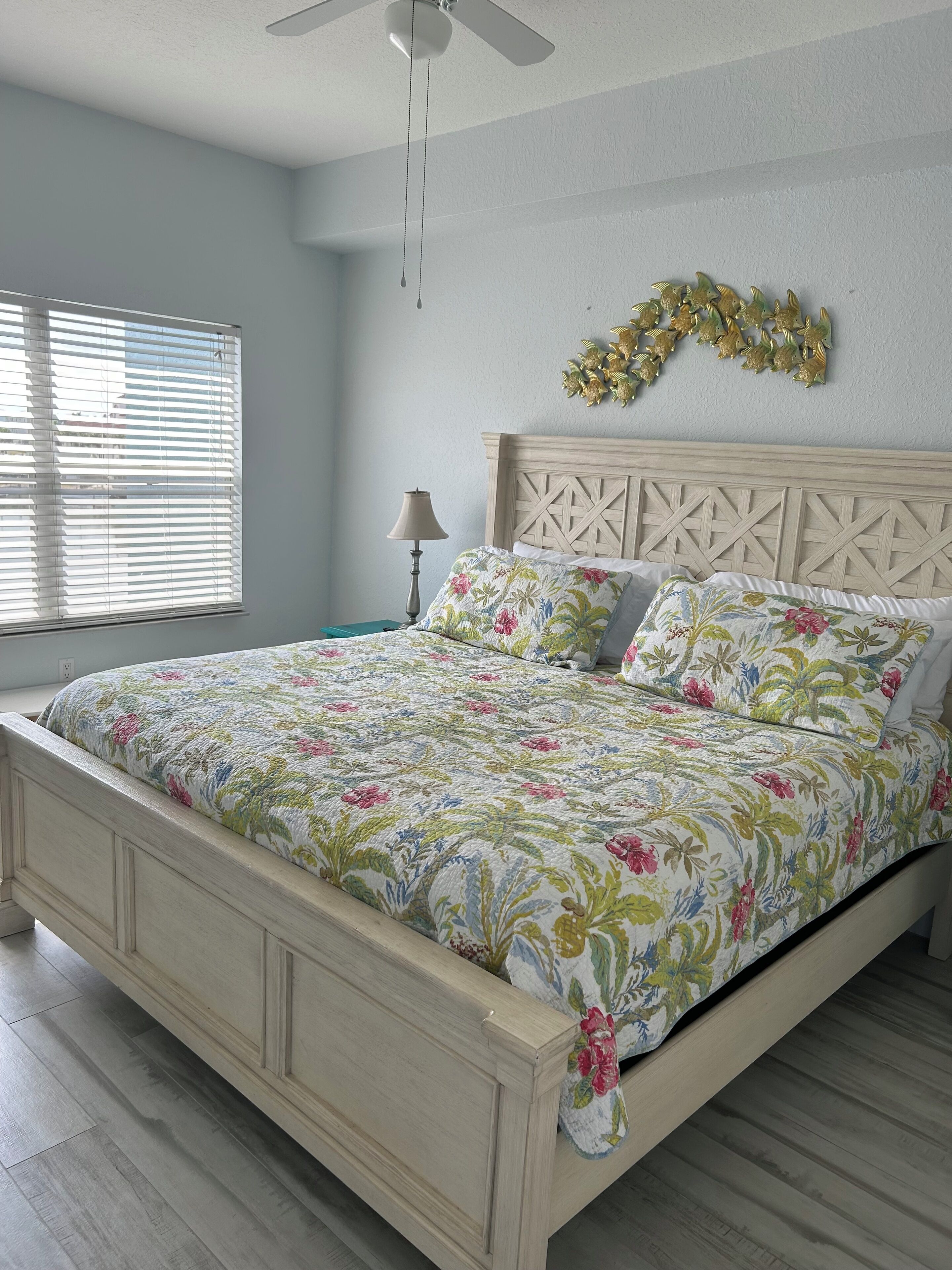 A bedroom with a bed, a ceiling fan, a window with blinds, a nightstand, and a wall decoration.