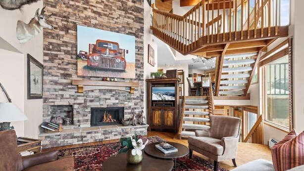 A living room with a stone fireplace, a wooden staircase, and a large window. - Abode at Highpoint, private hot tub, pool table, mountain retreat (Park City)