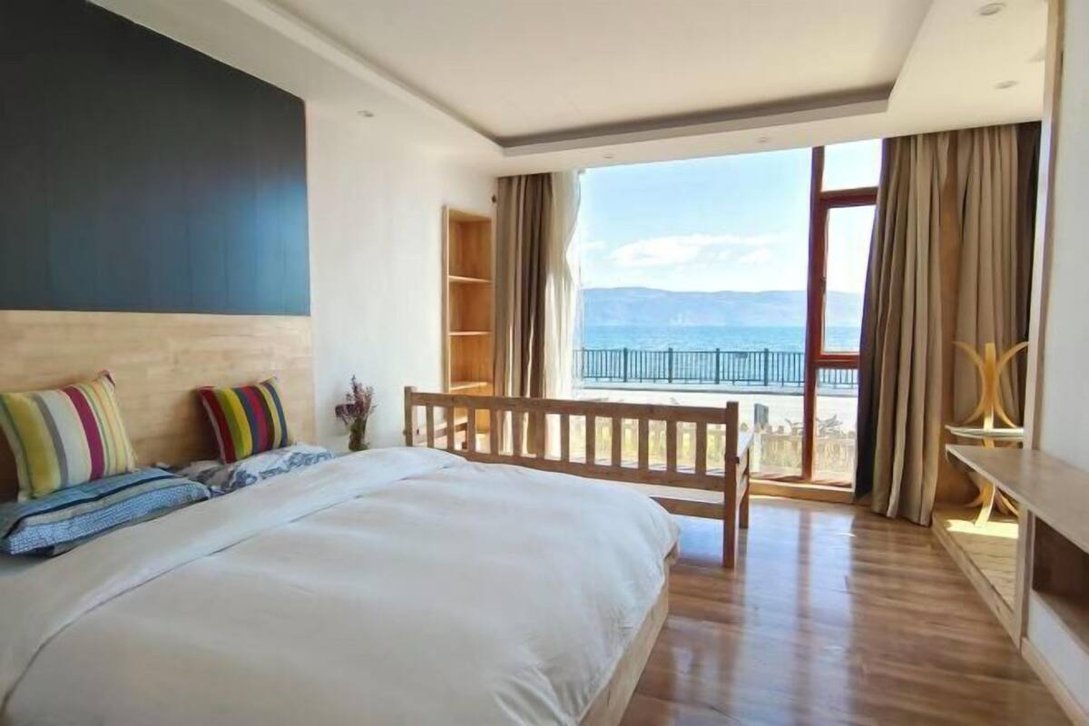 Une chambre avec un grand lit, un banc en bois, une bibliothèque et une vue sur la mer depuis un balcon.