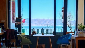 A room with a large window overlooking a body of water and mountains. There is a table with two chairs, a wooden floor, and a shelf with items on the left. - HillSea Green Holiday Inn , Dali (Dali City)