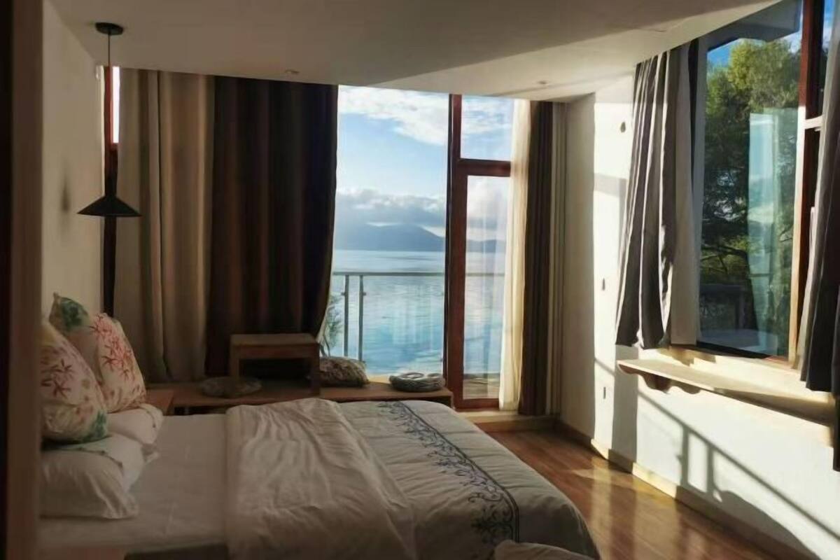 Une chambre à coucher avec un lit, une table de chevet, une lampe et une vue sur la mer grâce à de grandes fenêtres.