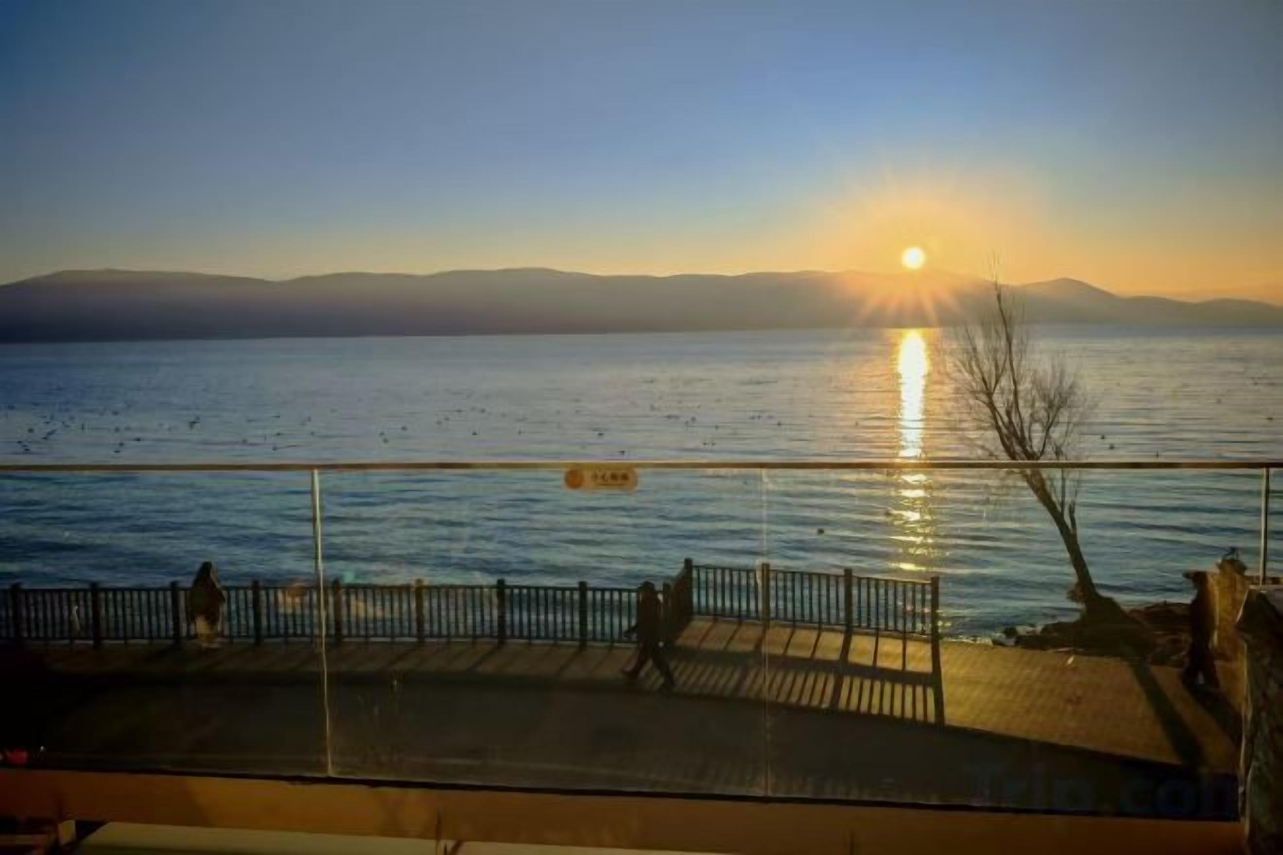 A sunset view from a balcony with a railing, a body of water, and distant mountains.