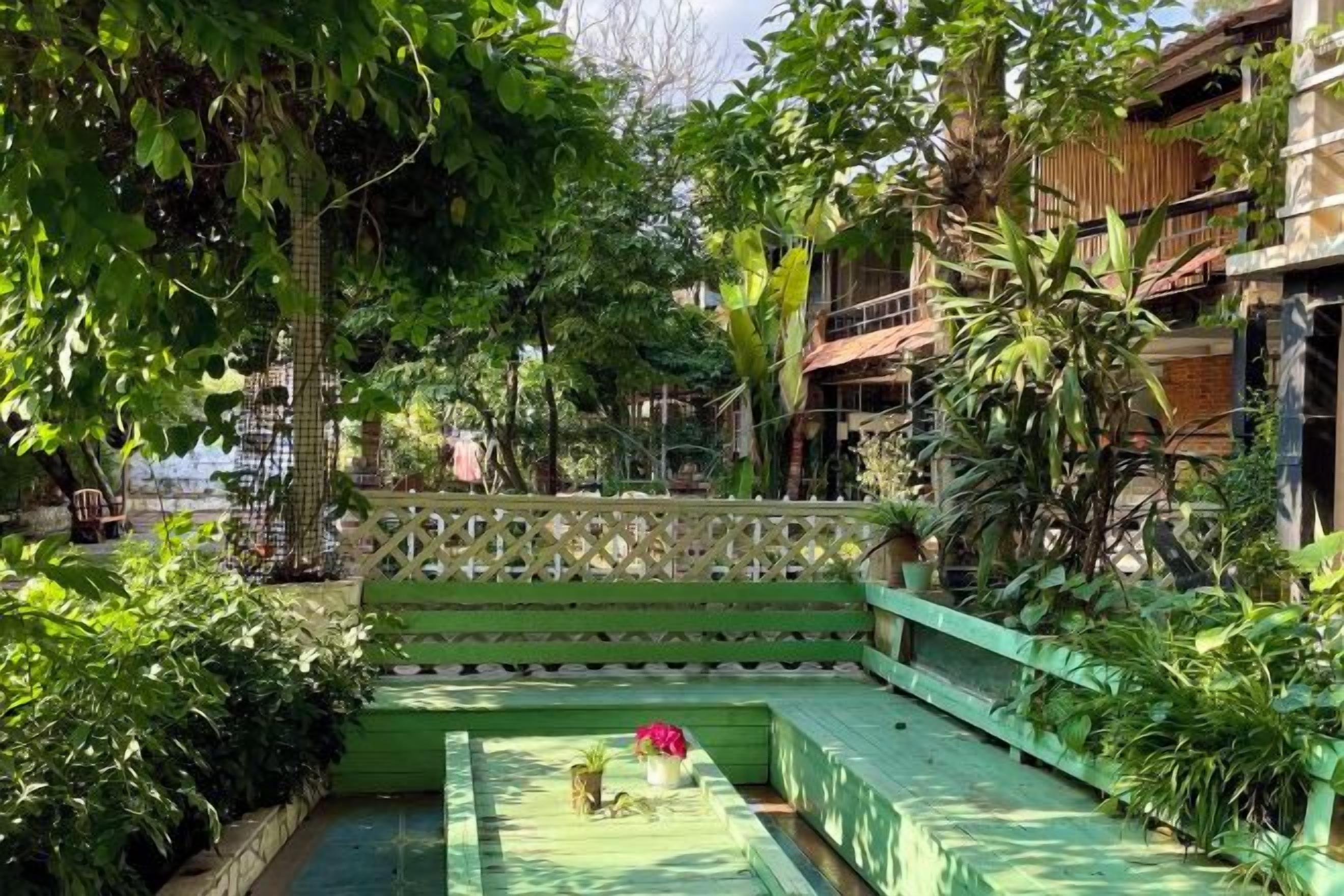 A green outdoor seating area with a bench, surrounded by lush greenery and a wooden fence.