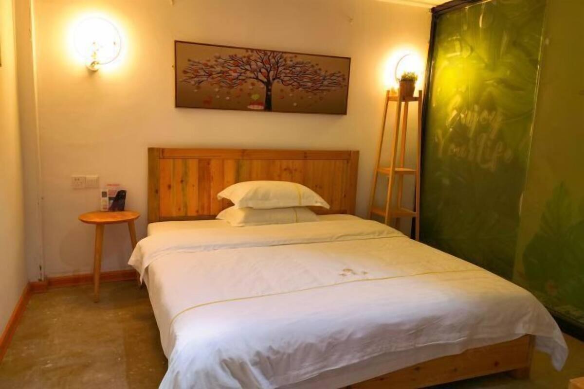 A single bed with a wooden headboard, a small bedside table, a wall-mounted light, and a painting of a tree.