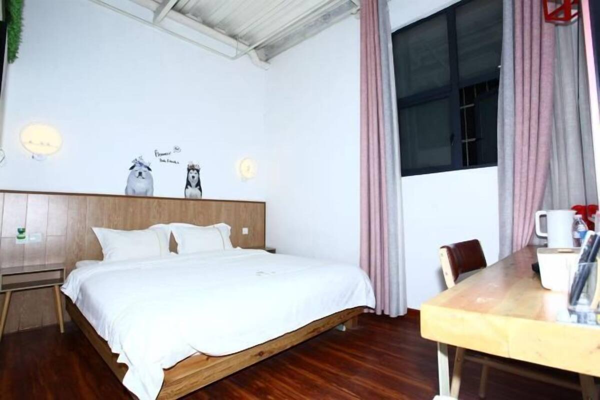 A bedroom with a wooden bed, a bedside table, a window with curtains, and a wooden dining table with chairs.