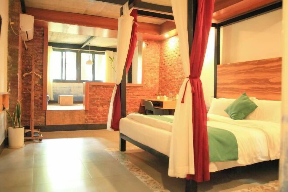 A bedroom with a bed, a desk, a chair, a potted plant, and a view of the outside.