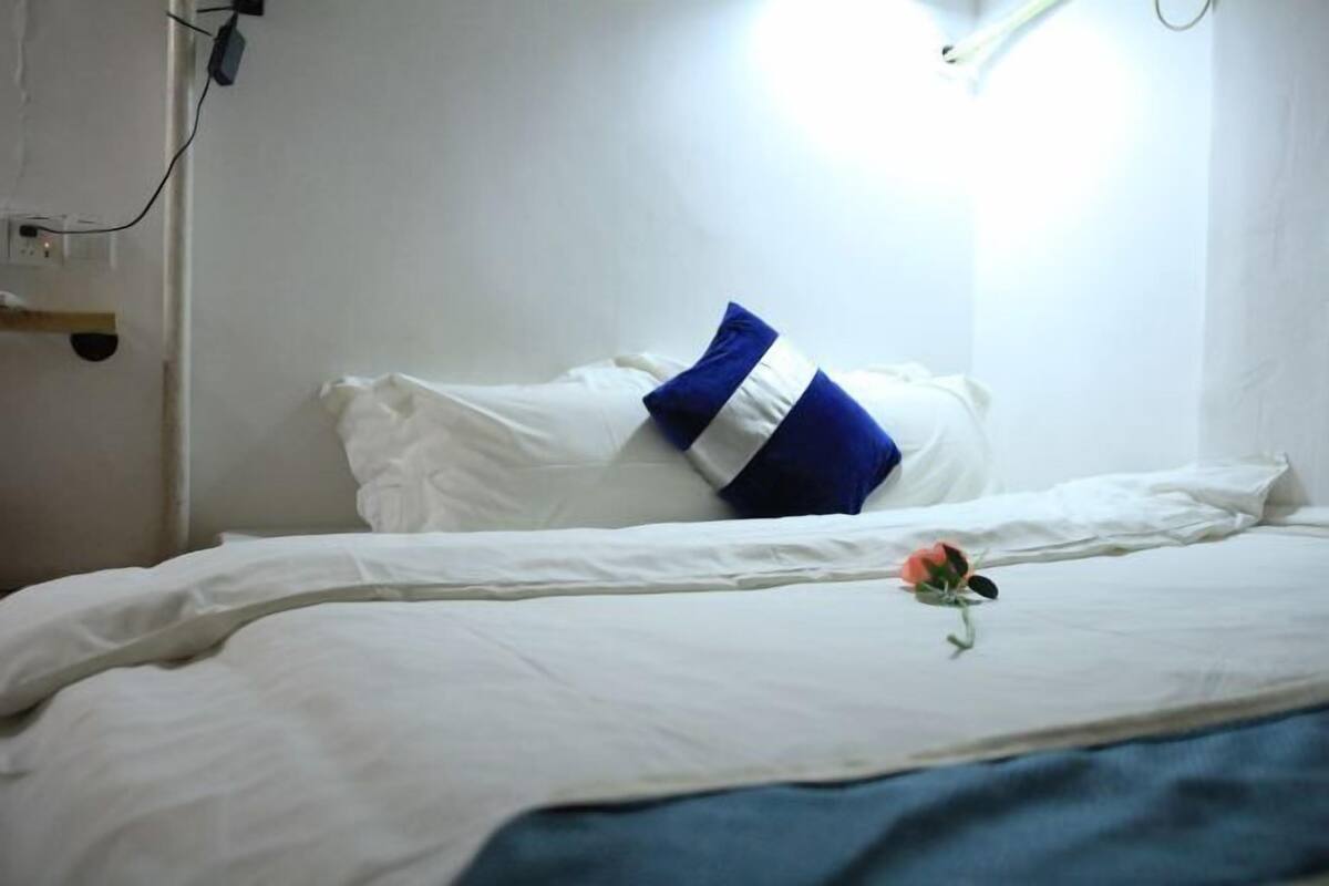 A bed with white bedding and a blue pillow, a single rose on the bed, and a white wall with a power outlet.