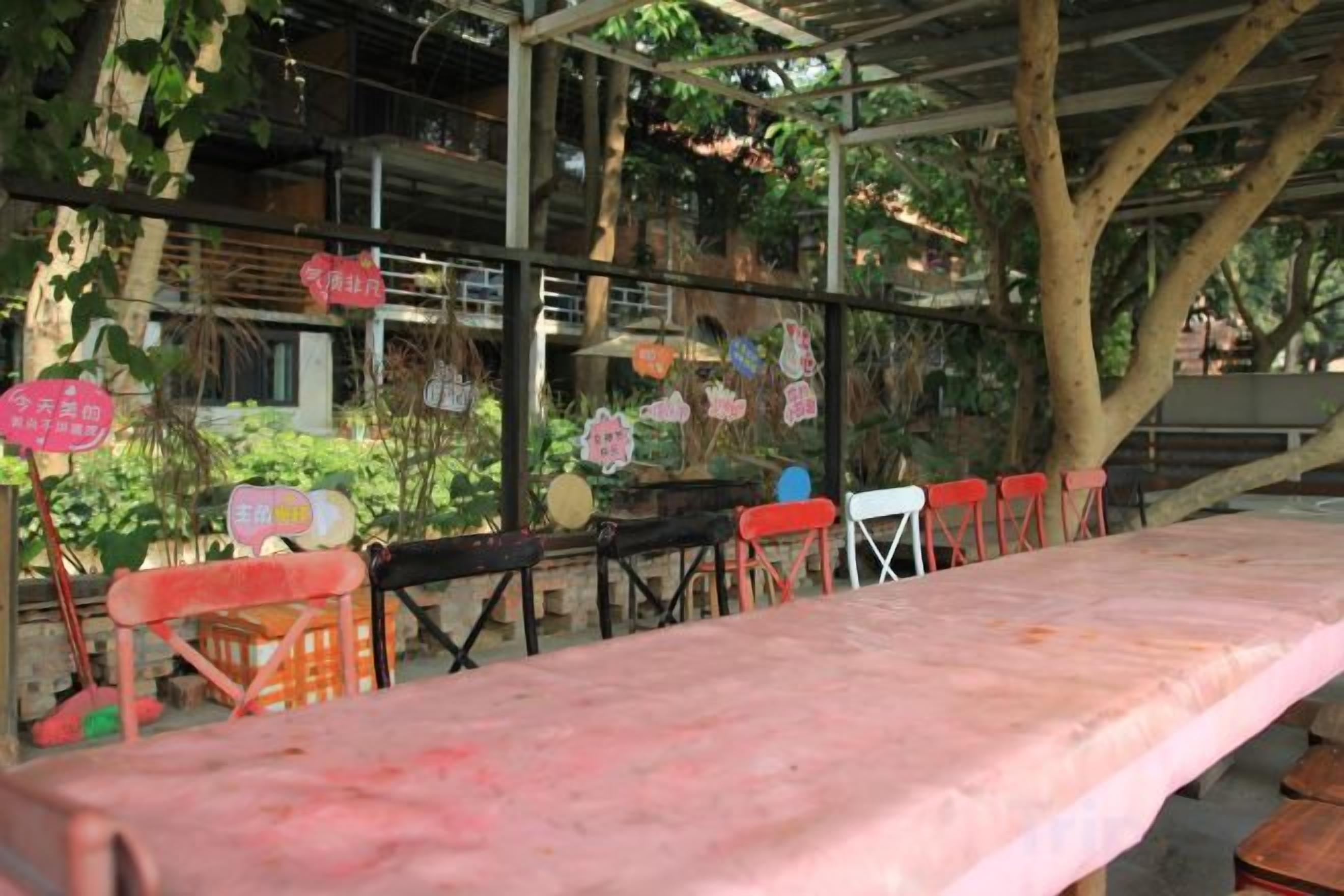 Un área de comedor al aire libre con una mesa larga y sillas, rodeada de árboles y una estructura de edificio.