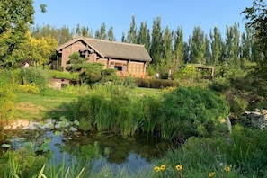 A rustic wooden house with a pond in front, surrounded by lush greenery and trees. - Goose city story (Baise)