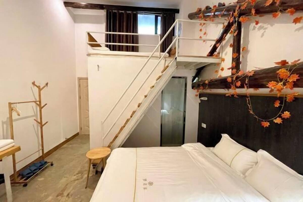 A bedroom with a bed, a wooden coat rack, a small stool, and a staircase leading to an upper level.