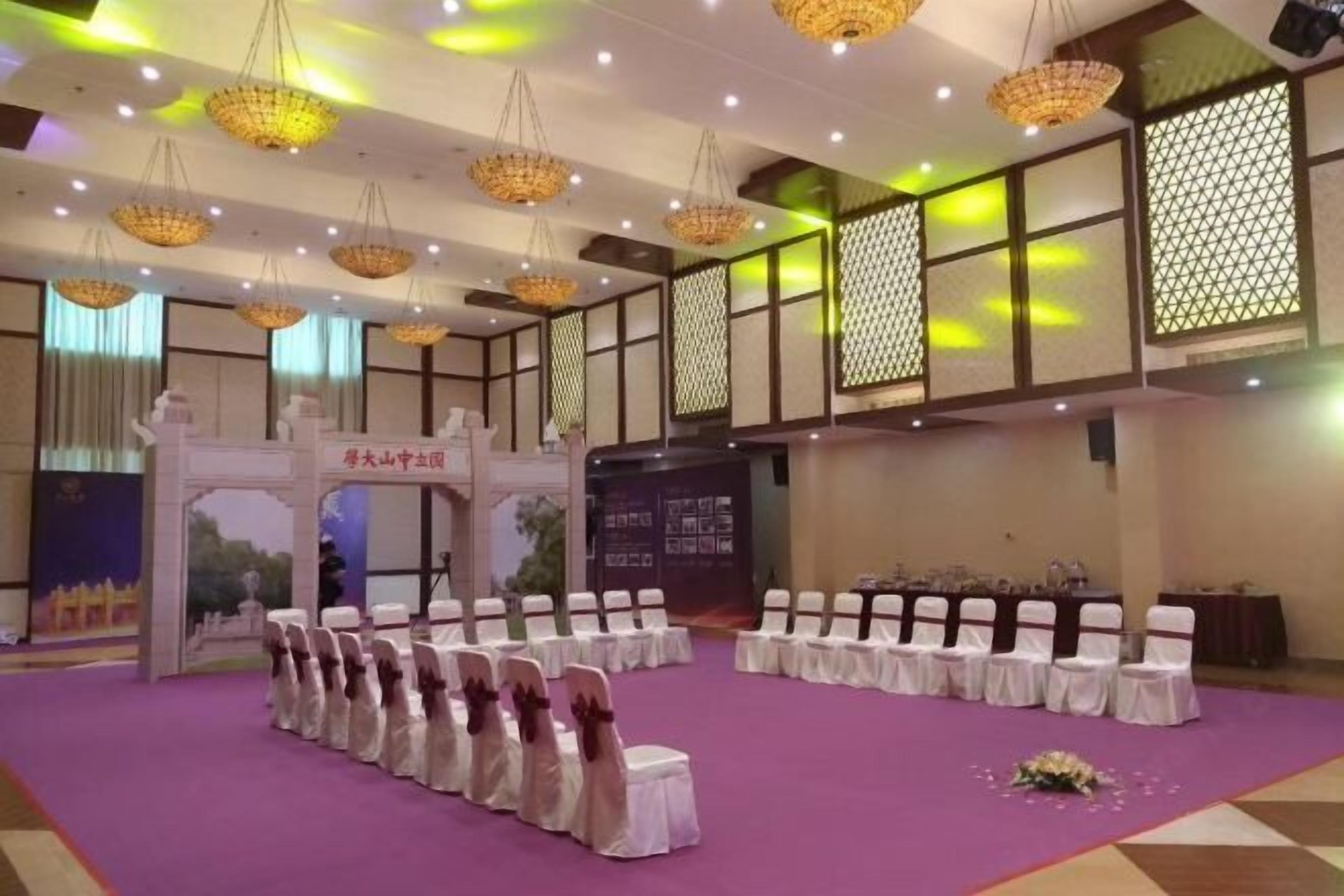 Un salón de banquetes con filas de mesas y sillas blancas, una alfombra púrpura e iluminación decorativa.