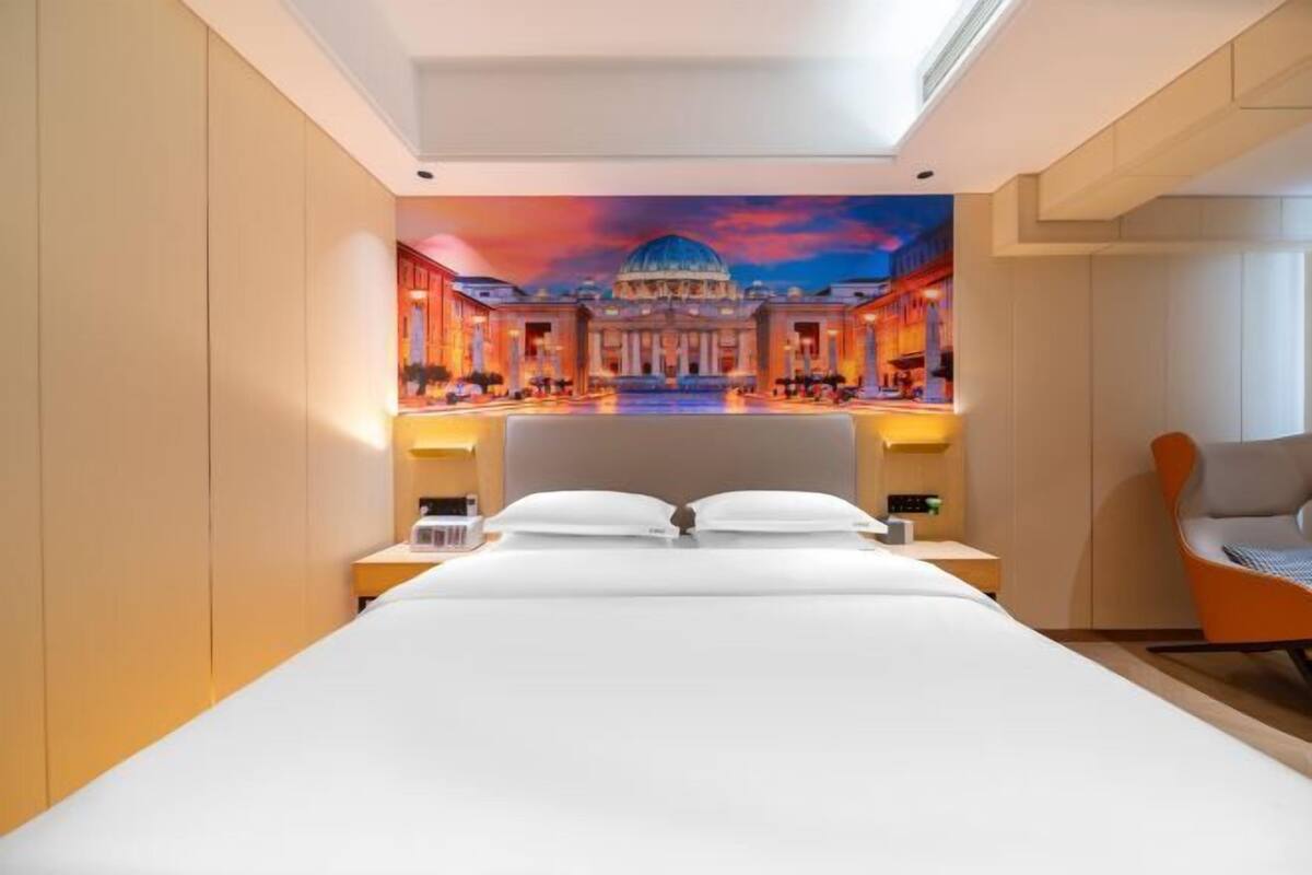 A hotel room with a large bed, two bedside tables, a nightstand, an orange chair, and a mural of a building on the wall.