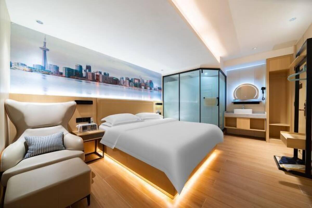 A modern hotel room with a large bed, a seating area with a chair, a glass-enclosed walk-in closet, and a cityscape mural on the wall.