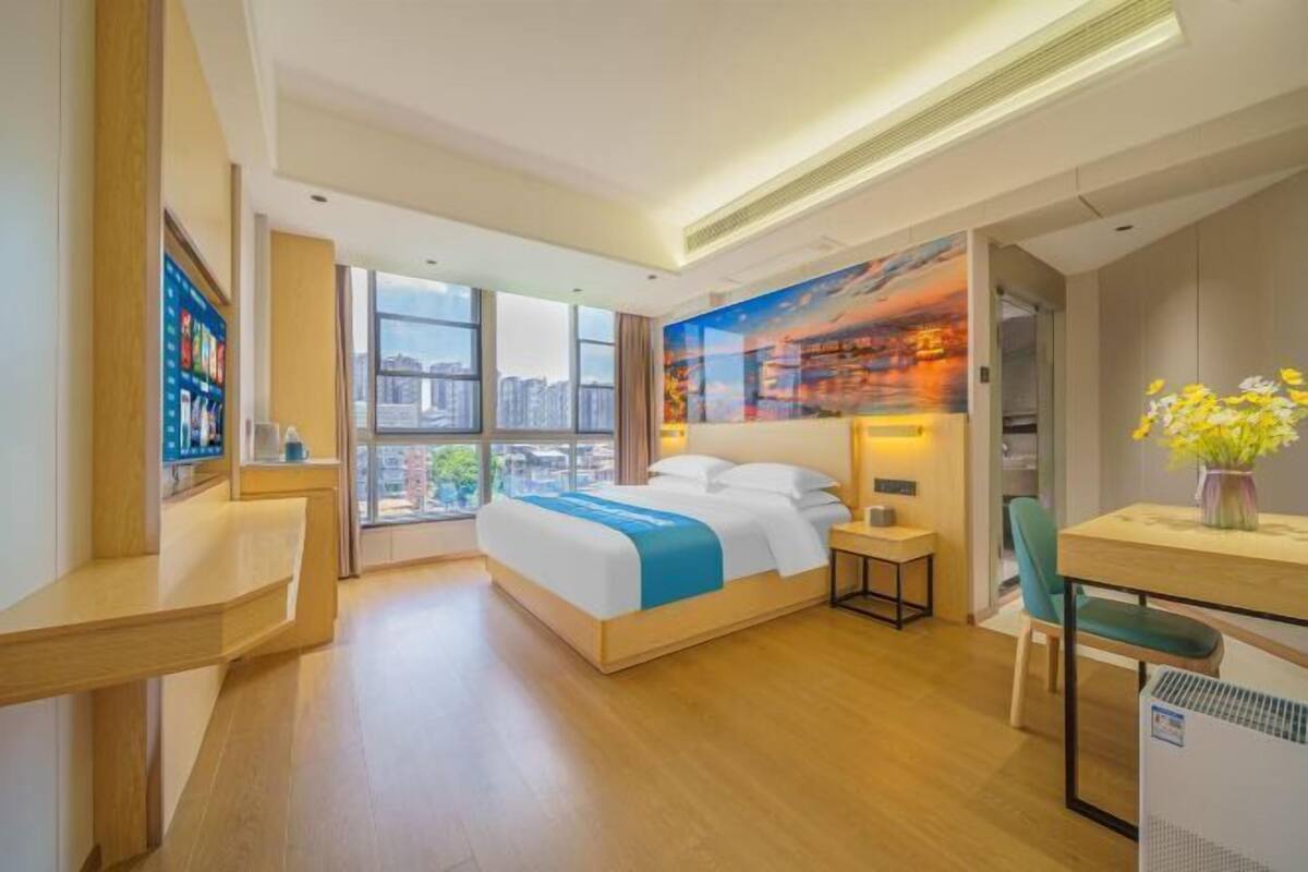 A modern hotel room with a large bed, a desk, a chair, and a view of the city through large windows.