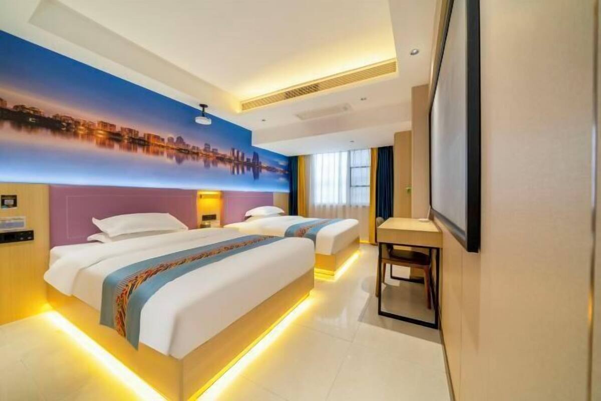 A hotel room with two beds, a cityscape mural, a desk, and a TV.