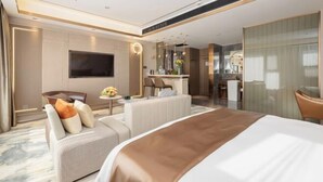 Een moderne hotelkamer met een groot bed, een zithoek met een bank en fauteuils, een eettafel, een televisie en een badkamer zichtbaar achter een glazen wand.