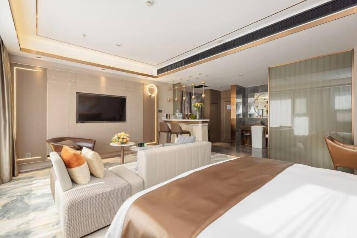 Una camera d'albergo moderna con un ampio letto, una zona salotto con divano e poltrone, un tavolo da pranzo, una televisione e un bagno visibile attraverso un pannello di vetro.