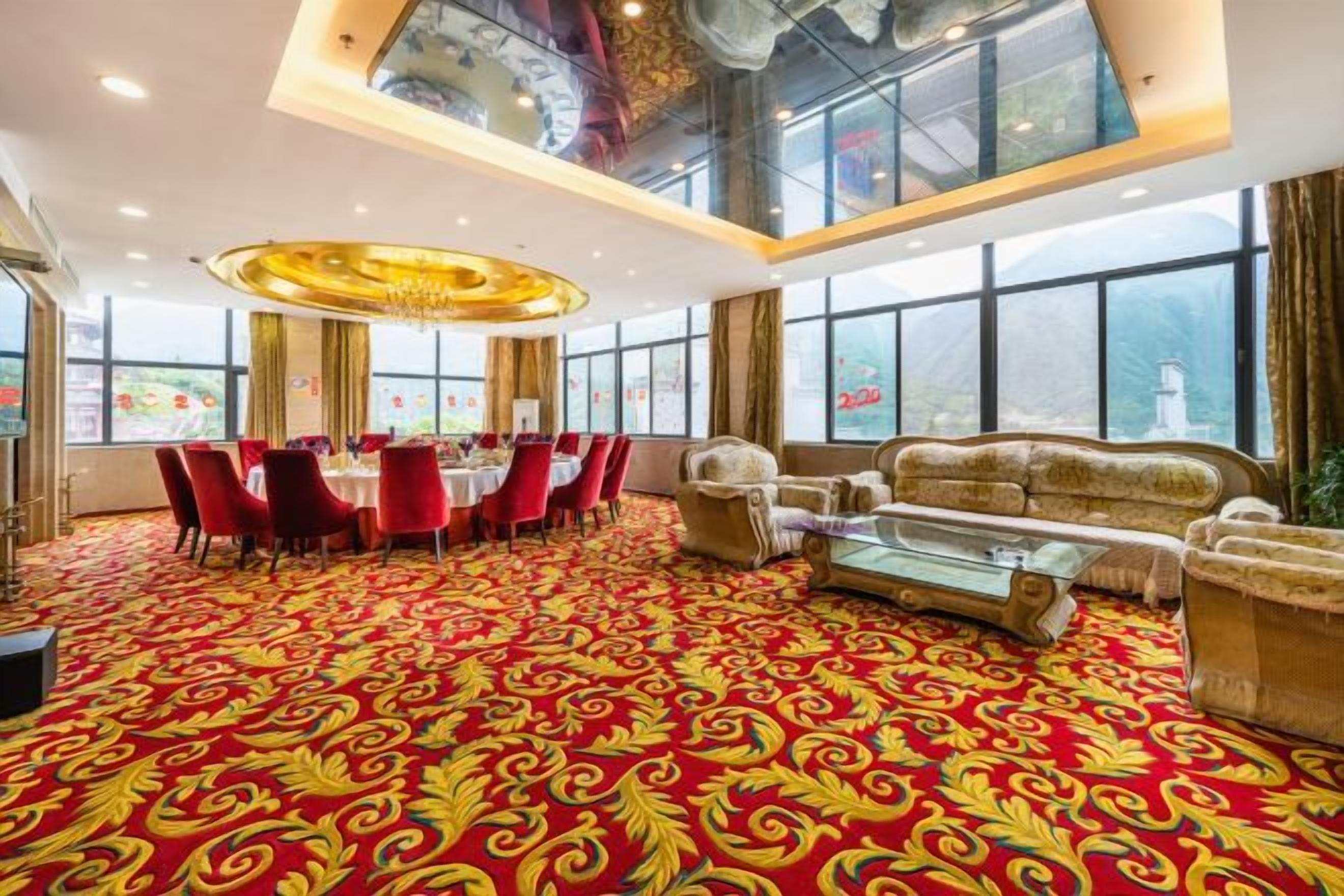 A hotel room with a large round table, red chairs, a sofa, and a patterned carpet.