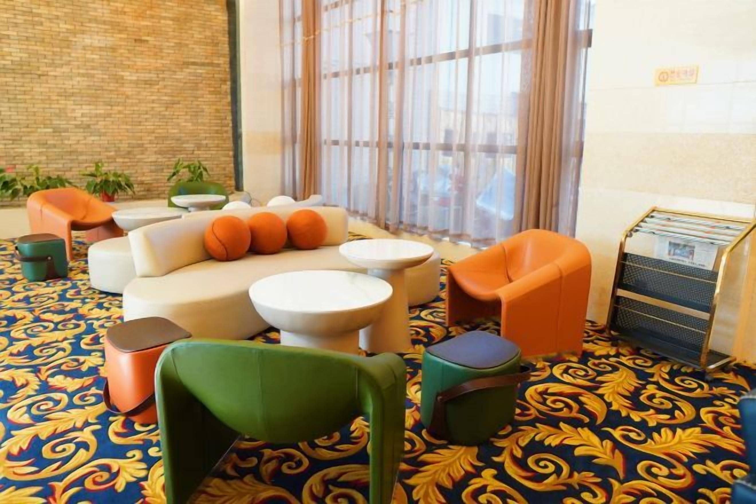 Un hall de hotel con una alfombra estampada, un sofá con cojines anaranjados, una silla verde y una pequeña mesa.