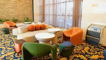 Un hall de hotel con una alfombra estampada, un sofá con cojines anaranjados, una silla verde y una pequeña mesa.