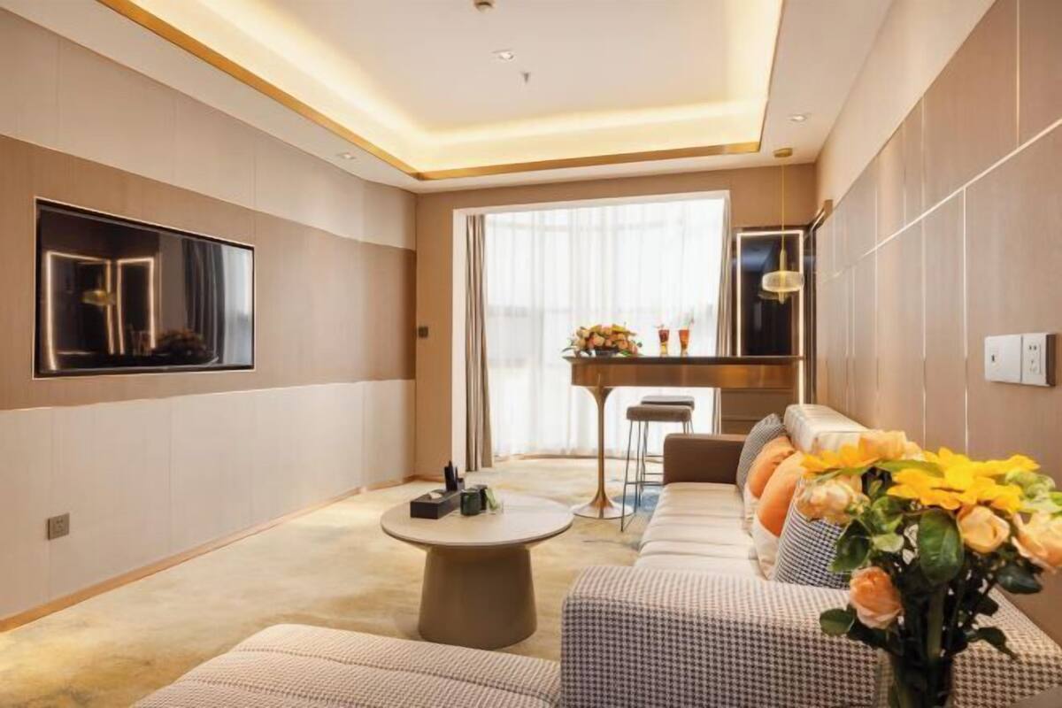Camera d'albergo moderna con una grande televisione a schermo piatto, un tavolino rotondo, un divano con cuscini decorati e un'area pranzo con un bancone e sedie.