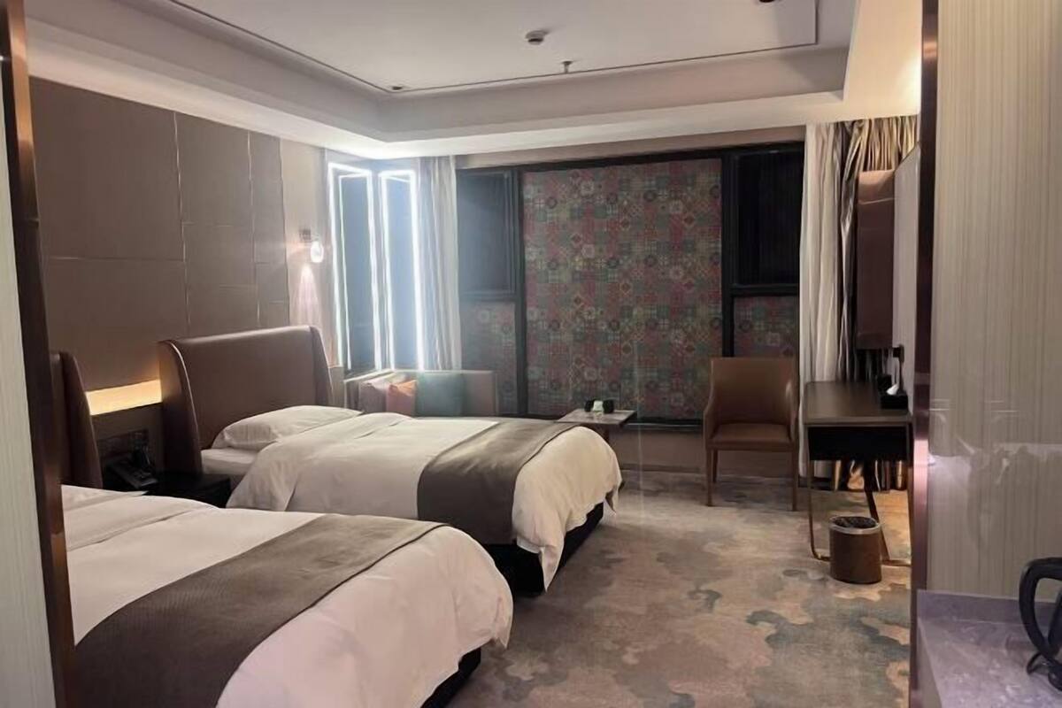Una camera d'albergo con due letti, una scrivania, una sedia e un'ampia finestra con tende.