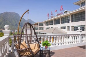 Un balcón de hotel con una silla de mimbre, una mesita con una planta y vistas del edificio del hotel y montañas al fondo.