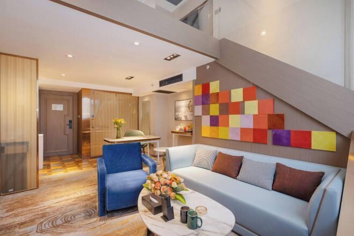Una camera d'albergo moderna con un divano, una poltrona blu, un tavolino da caffè con un vaso di fiori e un'opera d'arte astratta e colorata sulla parete.