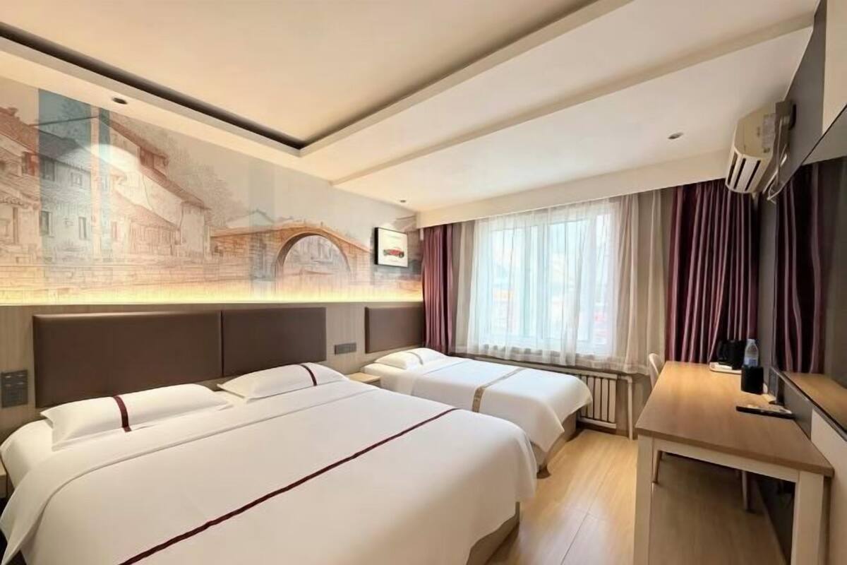 Ein Hotelzimmer mit zwei Betten, einem Schreibtisch, einem großen Fenster mit Vorhängen und einem Wandgemälde.
