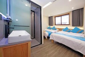Une chambre d’hôtel au design moderne, comprenant deux lits avec une literie bleue et blanche, une grande fenêtre avec des rideaux, un lavabo intégré et une cabine de douche en verre.