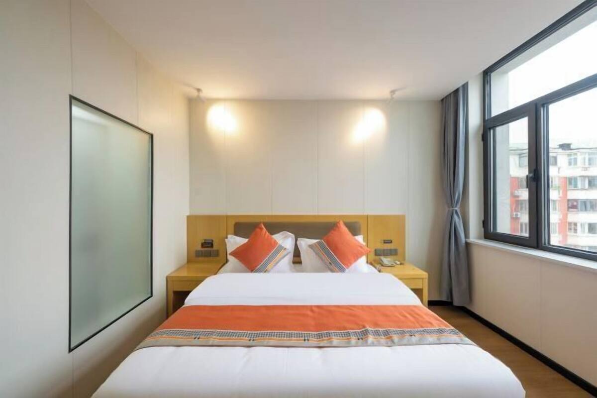 A hotel room with a large bed, two bedside tables, a nightstand, a large window with curtains, and a view of buildings outside.