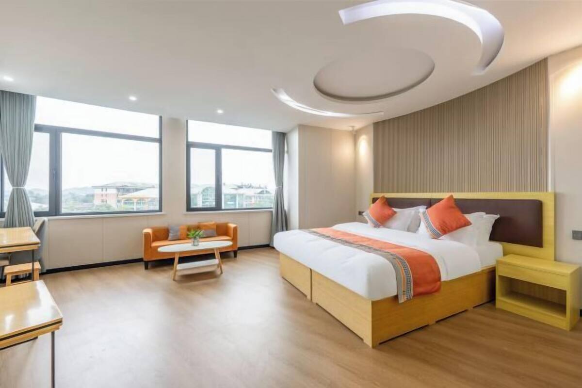 A hotel room with a large bed, a desk, a chair, a small table, and a view of the city through large windows.
